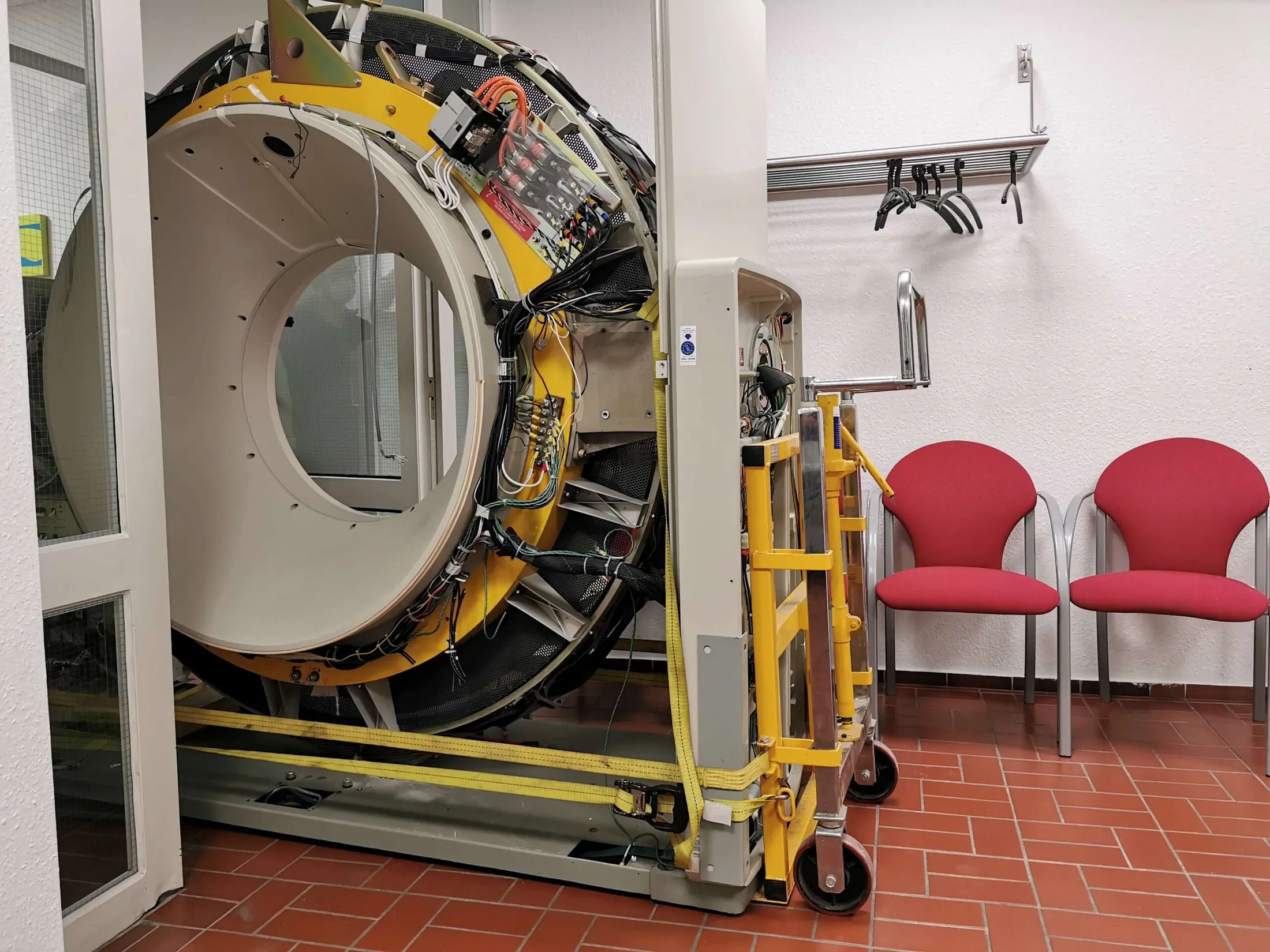 Geöffneter CT-Scanner mit sichtbarer Verkabelung auf Transportgestell neben roten Stühlen im Untersuchungsraum.